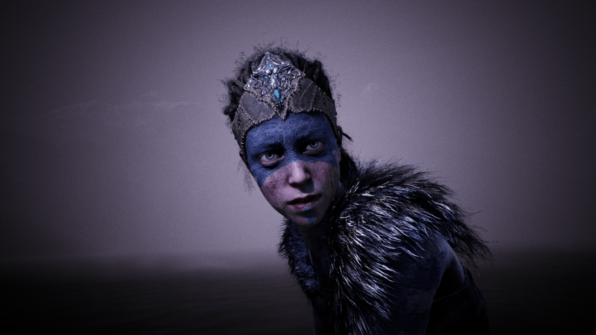A screenshot showing a medium close-up on a young woman with dark blue facepaint on the upper half of her face.  She's wearing a leather crown of sorts with jewels in the centre, as well as a short-sleeved tunic with a large fur collar.  She has a neutral expression.