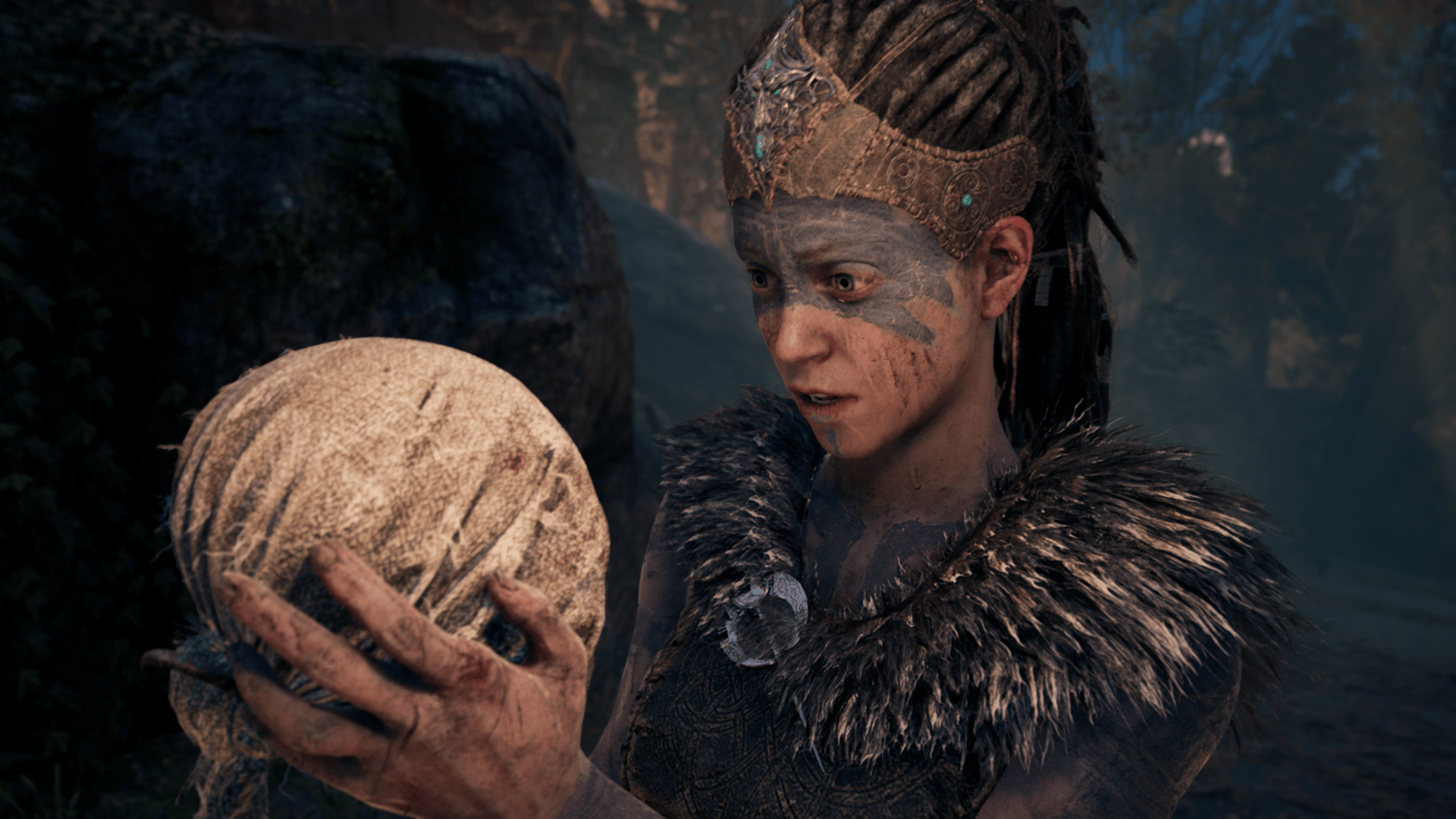 A screenshot showing a medium close-up on a young woman with dark blue facepaint on the upper half of her face.  She's wearing a leather crown of sorts with jewels in the centre, as well as a short-sleeved tunic with a large fur collar.  She is looking at a fabric-wrapped sphere that she's holding in her hands.